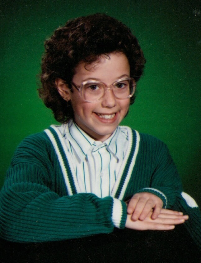 Kelley Shaw-Wade as young girl smiling for a school photo on a green background. She's on-brand, wearing a green striped button down shirt, a green cardigan and pink plastic glasses. Her curls are cut short, her hands are folded, and her dimples are showing.