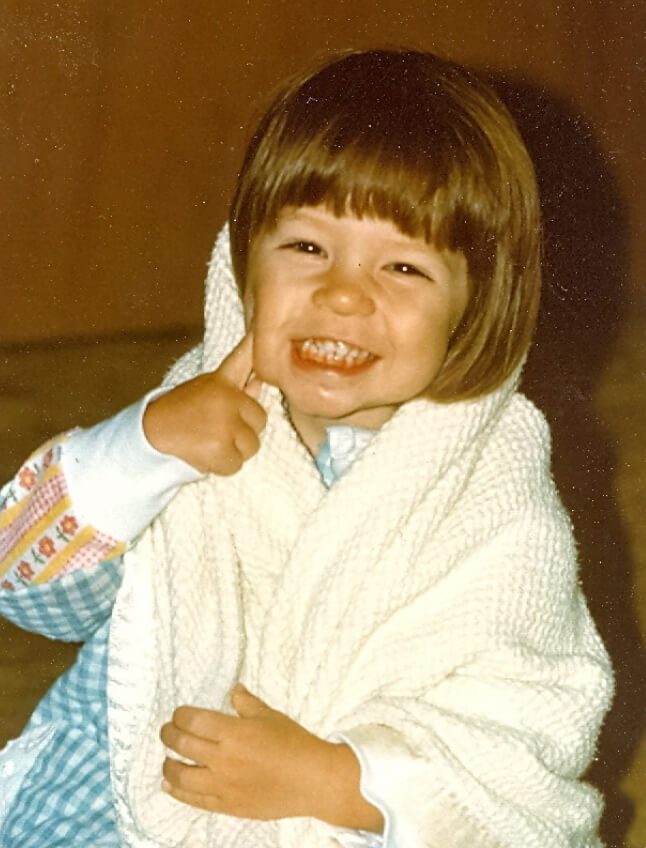 Kristine Wirth as a young child. She wears a top with flowers on the sleeve and a blue gingham dress, and is wrapped up in a waffle-knit blanket. She smiles broadly, with one finger poking her pudgy cheek.