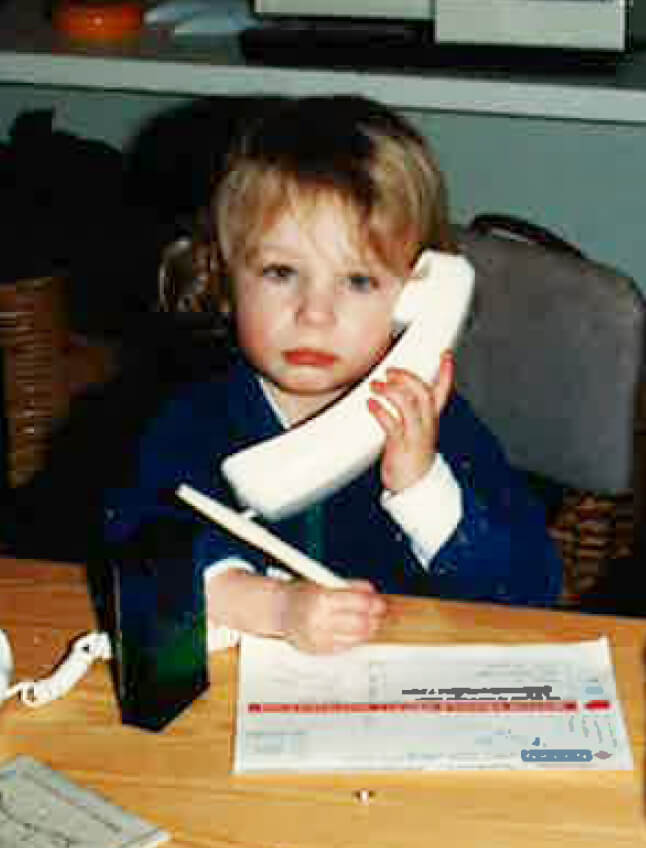 Leah Kasell as a young child seated at a desk. In one hand she holds a pen, poised to take notes; the other holds a corded phone up to her ear. She glances miserably at the camera.