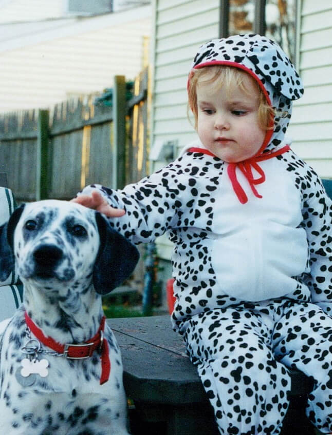 Rachel Suflita McKee as a young child dressed in a onesie dalmatian costume. Blonde curls peek out from a hood with red trim as she gently pets her mascot Luna, a real dalmatian beside her with a matching red collar.