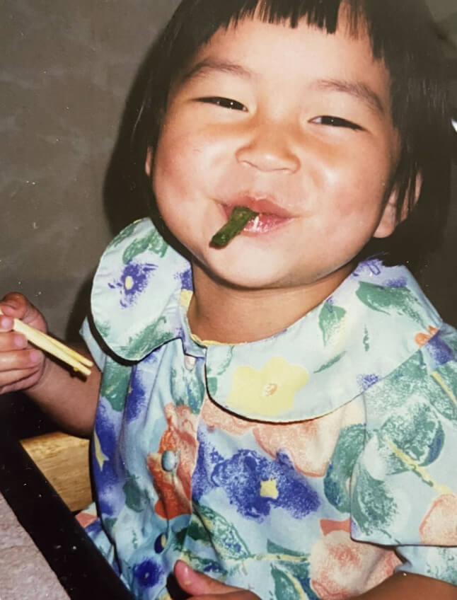 Susie Rowe Doan as a young child. She holds chopsticks, and even with a green bean hanging out of her mouth, she's smiling. She wears a floral top with a peter pan collar and she has both a bowl cut and hand-cut bangs.